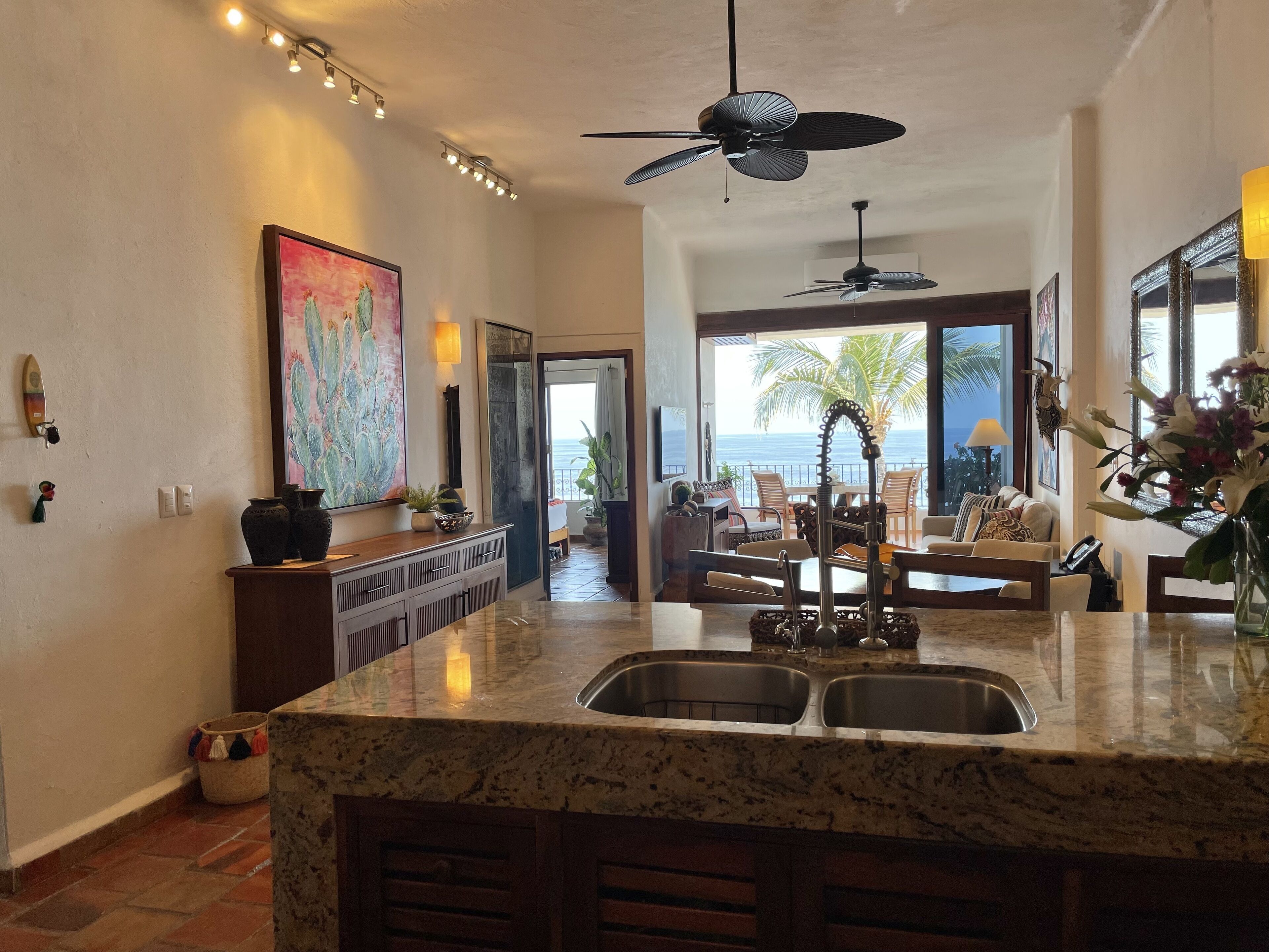 A kitchen with a marble countertop, two sinks, a ceiling fan, and a view of the ocean through large windows.