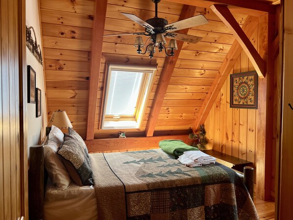 A cozy attic bedroom with wooden walls, a sloped ceiling, a bed with a quilt, a small window, and a ceiling fan. - Beautiful Timber Frame on Saco River enjoy Fall Foliage Near North Conway (Brownfield)