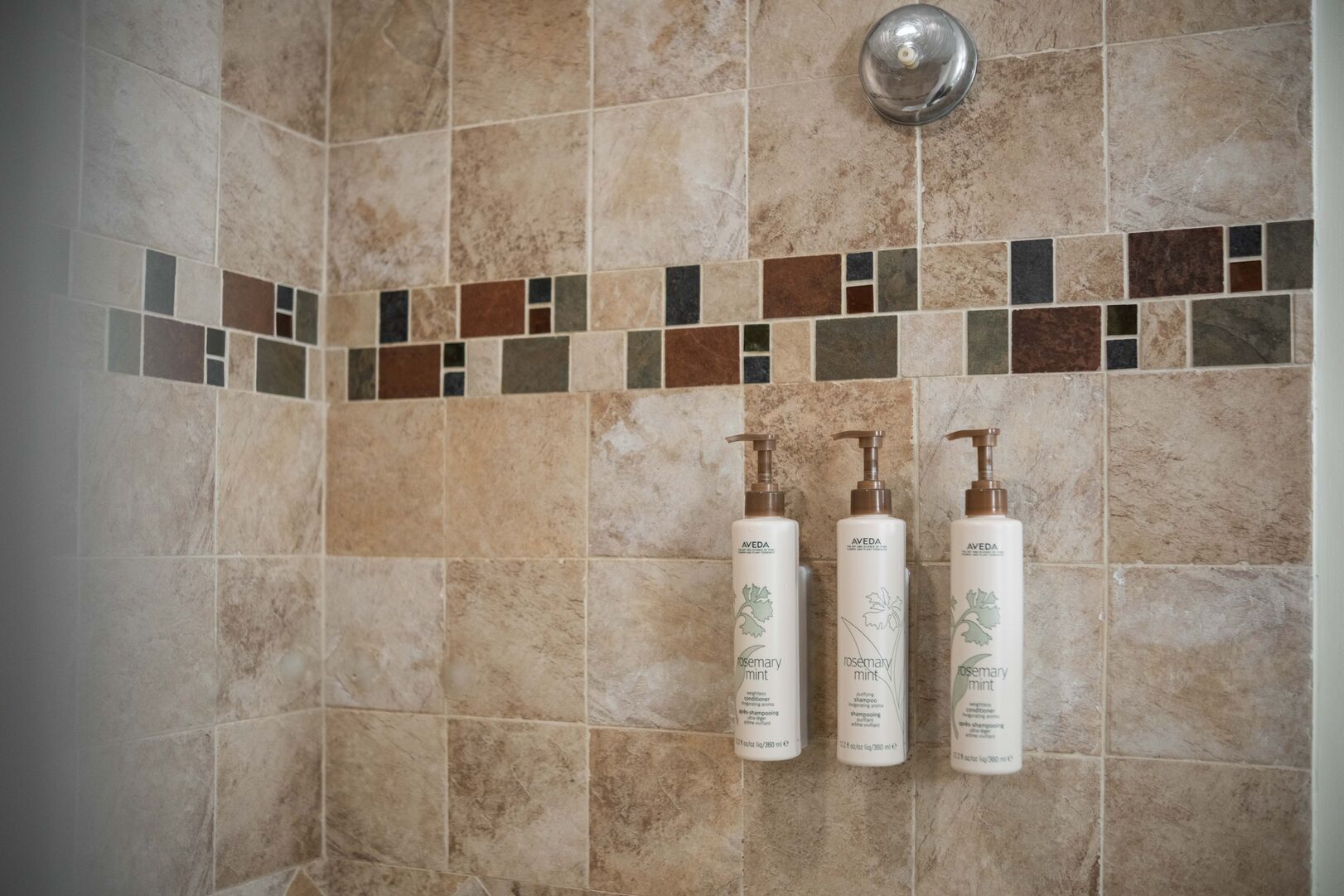 A bathroom with beige tiles and a mosaic border. There are three bottles of Aveda Rosemary Mint shampoo and conditioner on the wall.