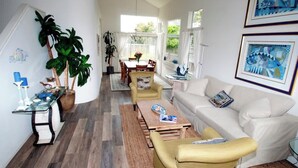 A spacious living room with a white sofa, a yellow armchair, and a coffee table. There is a dining area with a table and chairs, and a large window providing natural light. - Serenity at Natural Bridges  - 5 min Walk to Beach (Santa Cruz)