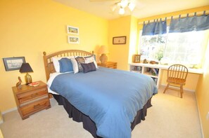 A bedroom with a bed, a nightstand, a chair, a window with curtains, and a bookshelf.