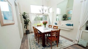 A dining area with a wooden table, chairs, and a patterned rug. There is a large window with curtains, a potted plant, and a mirror above the table. - Serenity at Natural Bridges  - 5 min Walk to Beach (Santa Cruz)