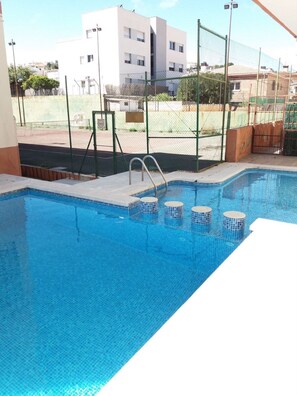 A swimming pool with steps leading into it, surrounded by a fence and buildings in the background.