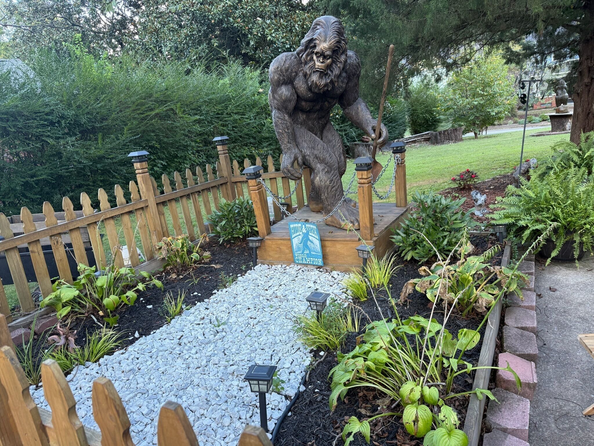 A garden with a wooden fence, a statue of a gorilla, and a sign that reads 'HIDE & SEEK WORLD CHAMPION'.