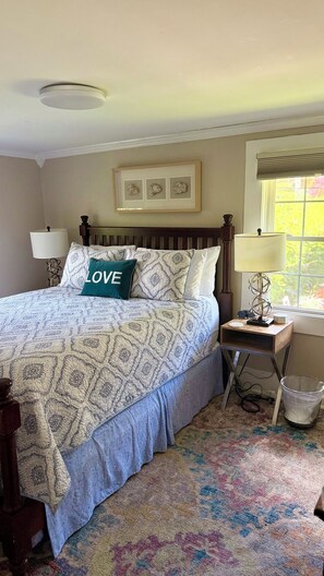 A bedroom with a bed, a bedside table with a lamp, a window with blinds, and a wall-mounted picture.