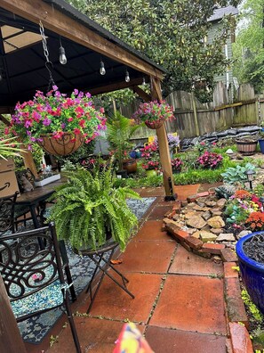 Un patio jardín con cestas de flores colgantes, un camino de ladrillo y diversas plantas en macetas.