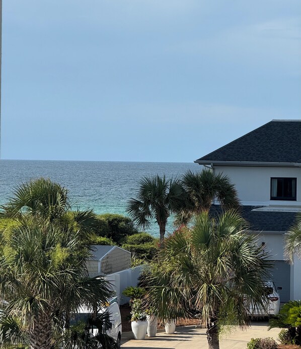 A coastal house with a view of the sea, surrounded by palm trees and a white building.