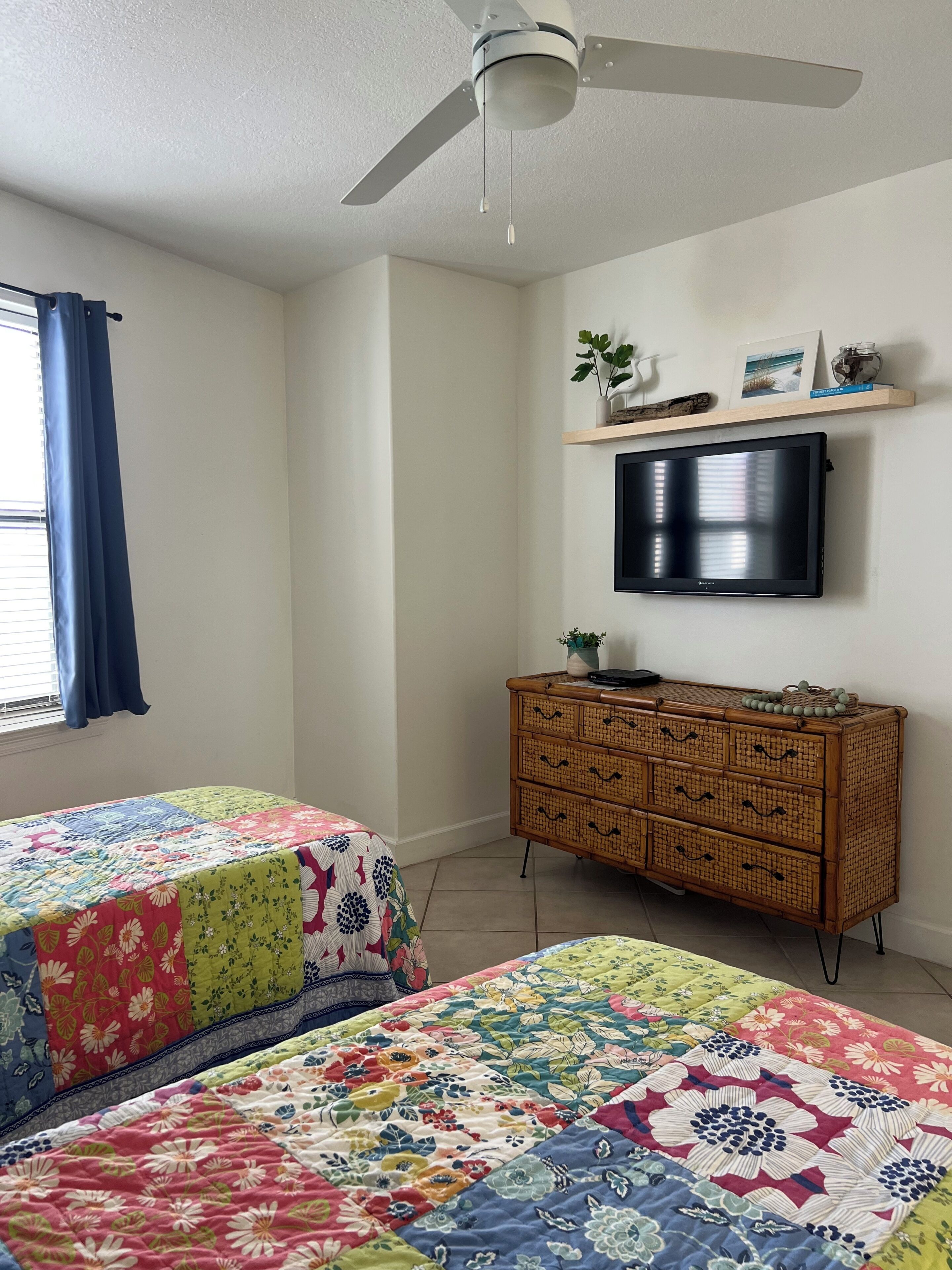 Une chambre avec deux lits, un ventilateur de plafond, un téléviseur posé sur une étagère et une fenêtre avec des rideaux.