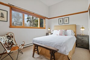 A bedroom with a large bed, a wooden bench, a chair, a nightstand, and a window with blinds.