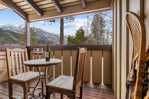 A wooden deck with a table and chairs, a bottle of wine, and a view of snow-capped mountains.