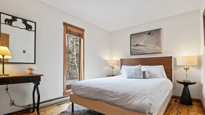 Een slaapkamer met een groot bed, een houten vloer, een bureau met een lamp en een raam met uitzicht op bomen.