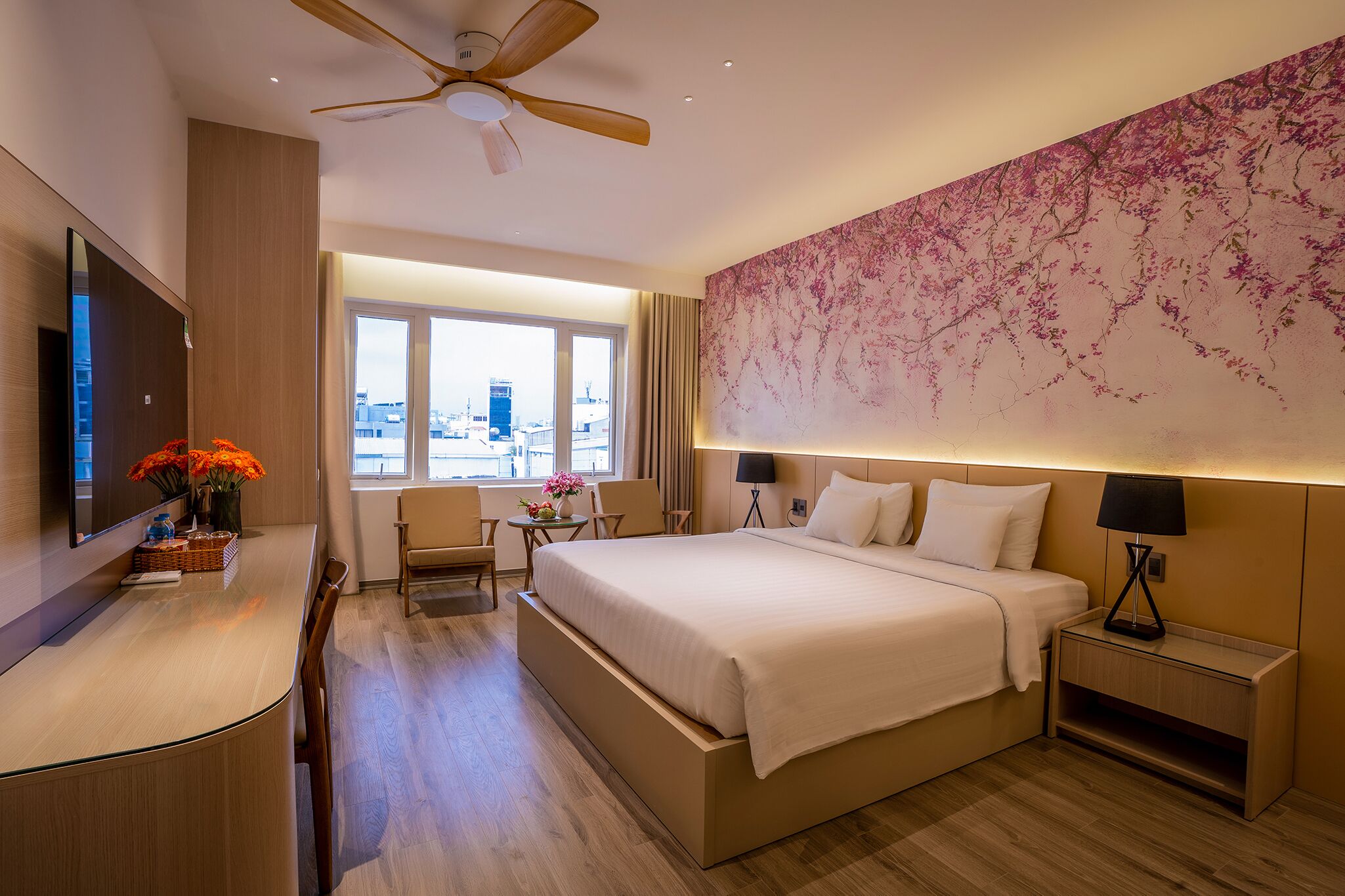 A modern hotel room with a large bed, a desk, a chair, a ceiling fan, a large window with a city view, and a wall-mounted TV.