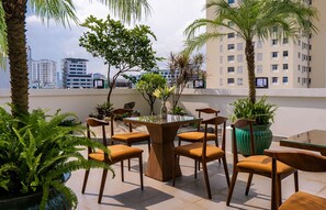 An outdoor seating area with wooden chairs and a glass table, surrounded by potted plants and palm trees, overlooking a cityscape. - Palago Hotel, Binh Thanh District (Ho Chi Minh City)