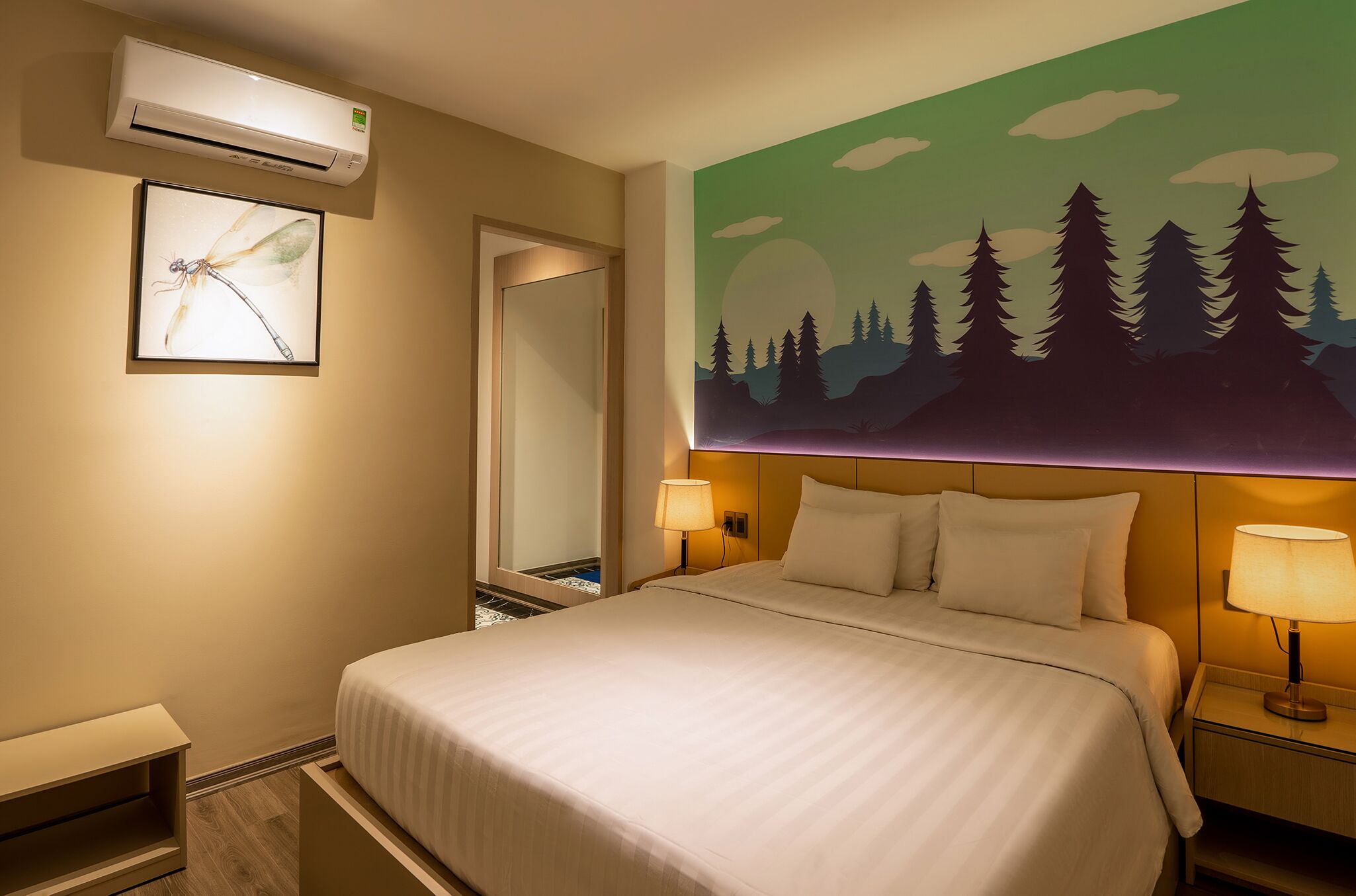 Hotelkamer met een bed, nachtkastje met lamp, airconditioning, lijst met libelle en muurschildering van bomen en een zonsondergang.