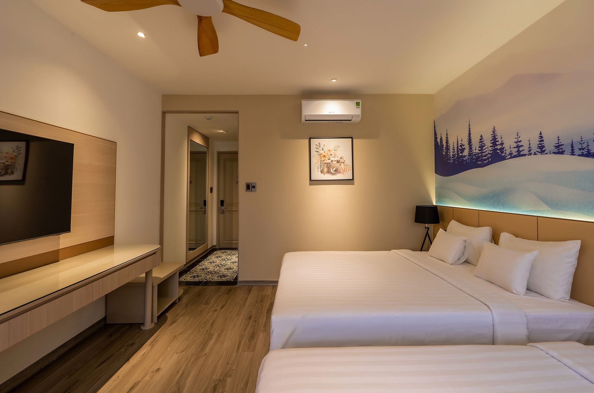 A hotel room with a bed, a TV, a ceiling fan, an air conditioner, and a painting of a snowy landscape.