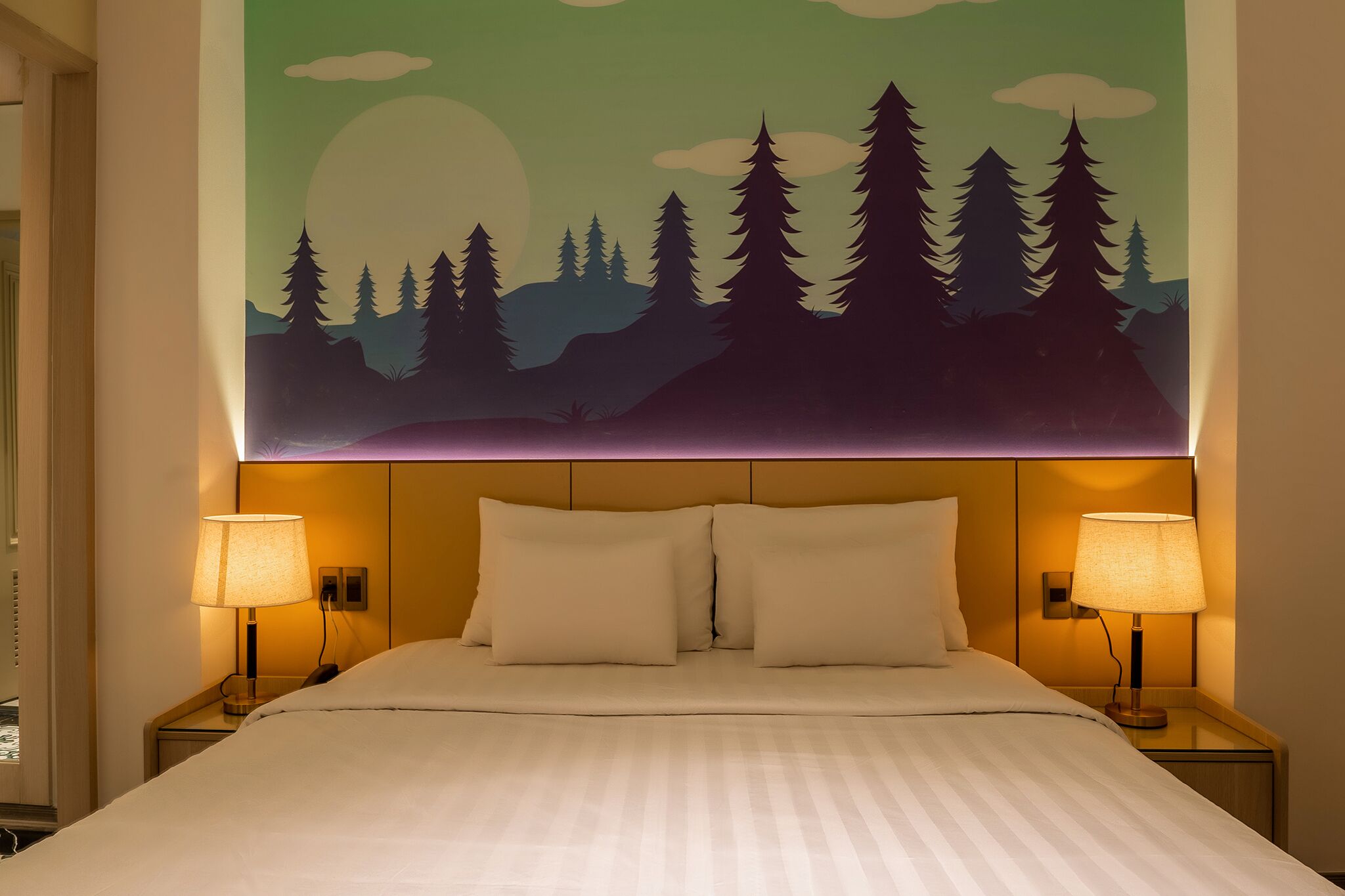 A hotel room with a bed, two bedside lamps, and a wall mural of a forest scene.