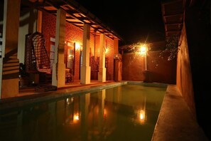 A nighttime swimming pool area with a brick building, a covered seating area, and a hanging chair. - Plantation Retreat (Pathadumbara)