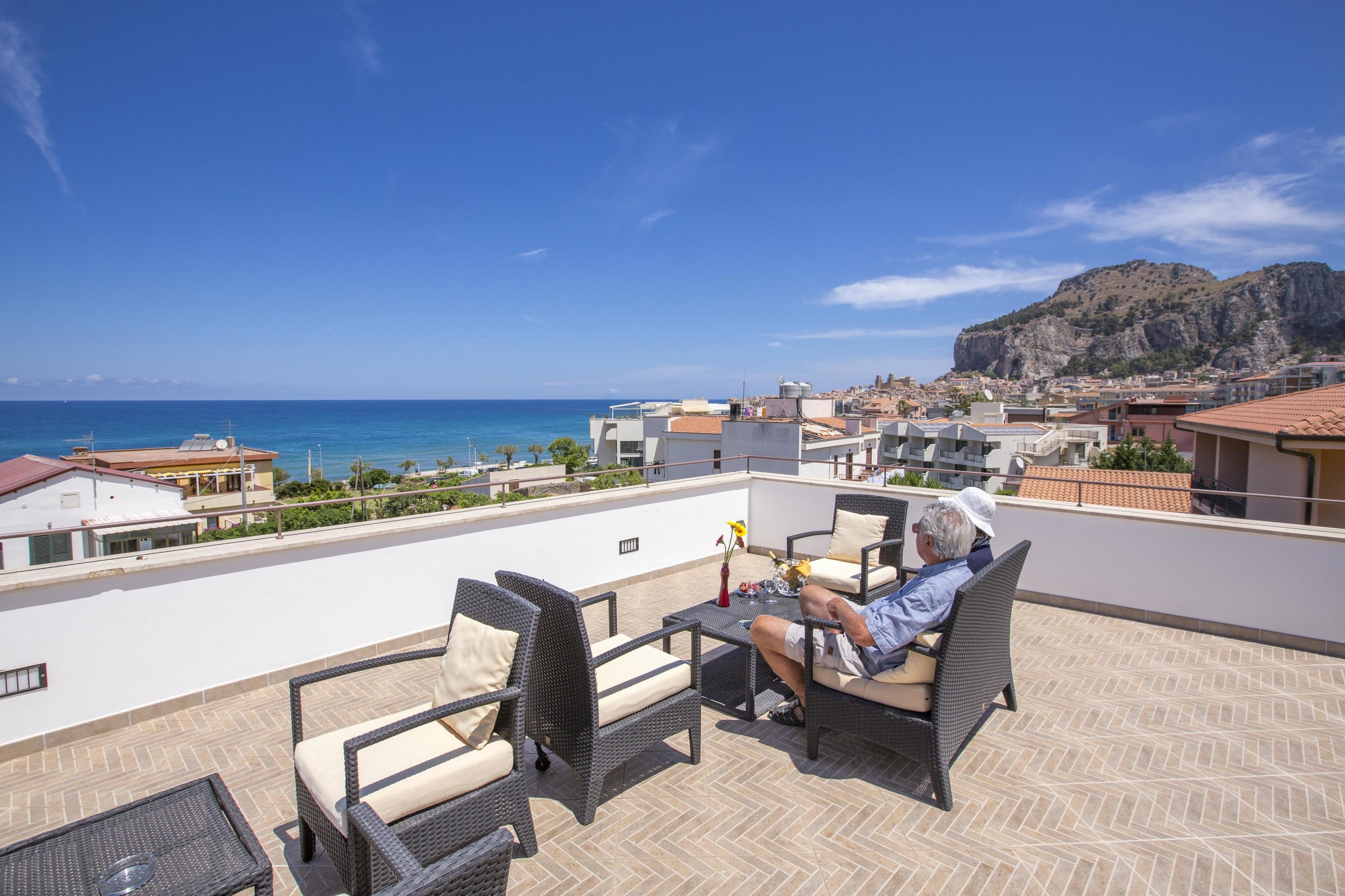 A rooftop terrace with outdoor furniture, a view of the sea, and a mountainous backdrop.