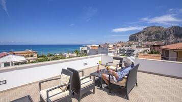 A rooftop terrace with outdoor furniture, a view of the sea, and a mountainous backdrop.