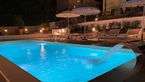 A swimming pool with a person diving, surrounded by lounge chairs and umbrellas, and well-lit by ambient lighting.