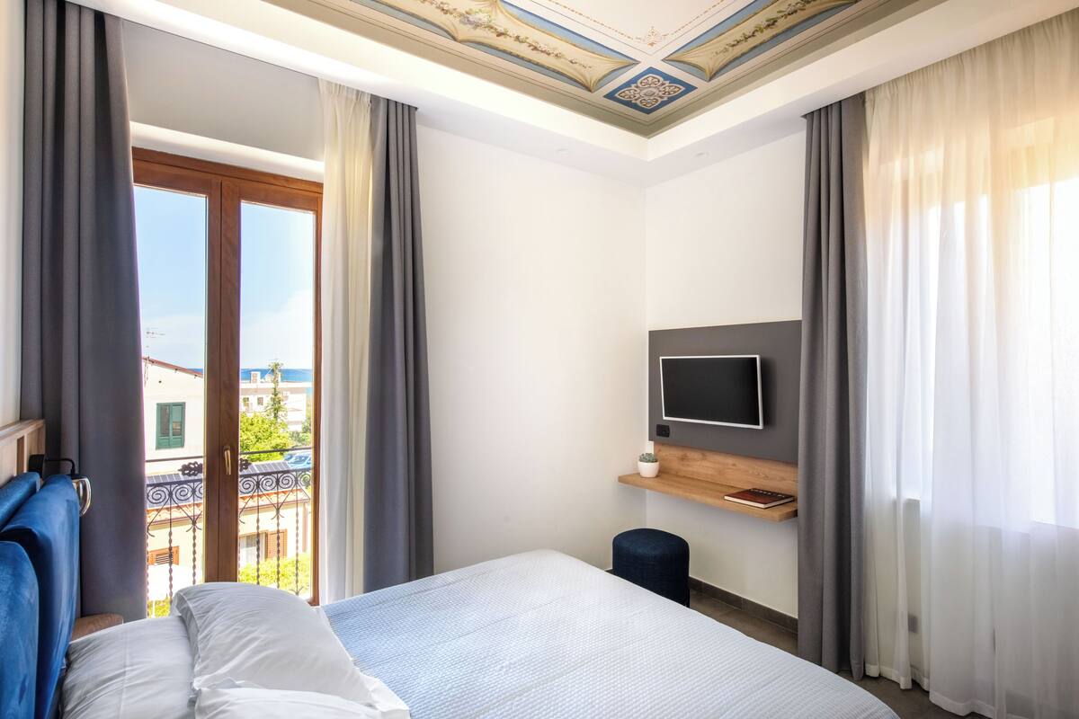 A hotel room with a bed, a TV mounted on the wall, a small stool, and a balcony with curtains.