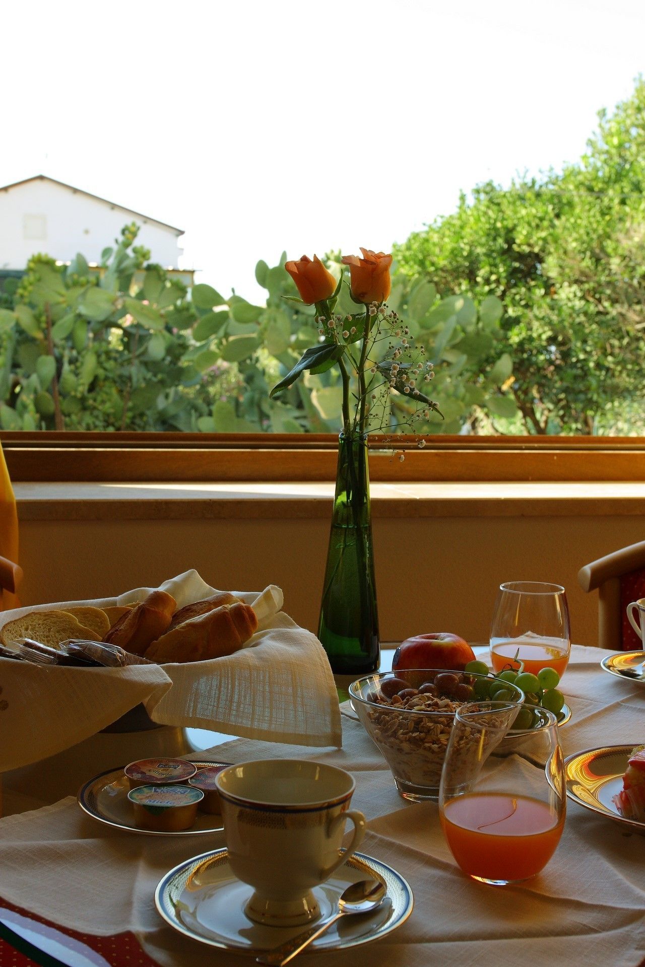 A breakfast table with croissants, a vase with flowers, and a view of greenery outside.