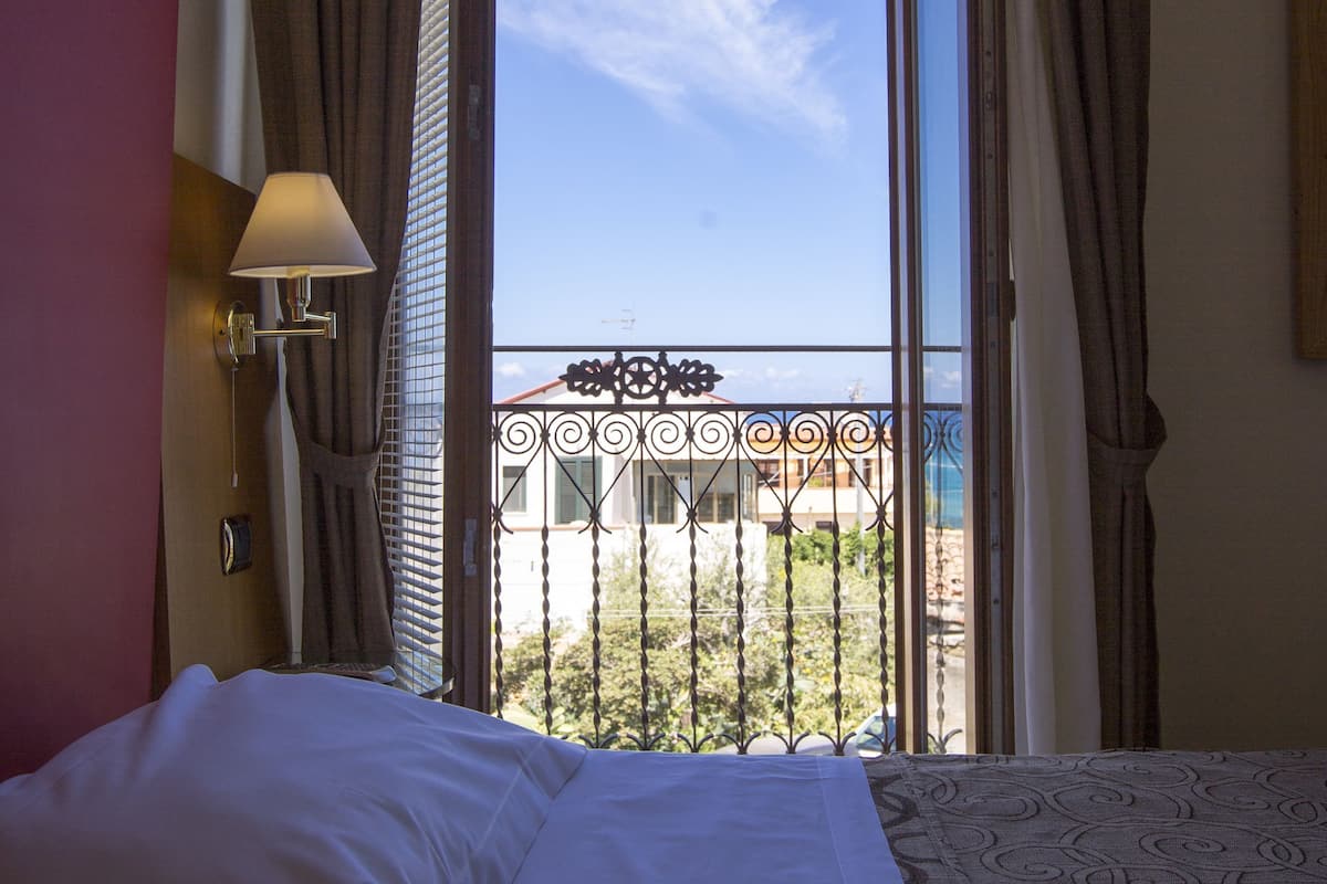A hotel room with a bed, a window with curtains, a view of the outside, and a balcony with a railing.
