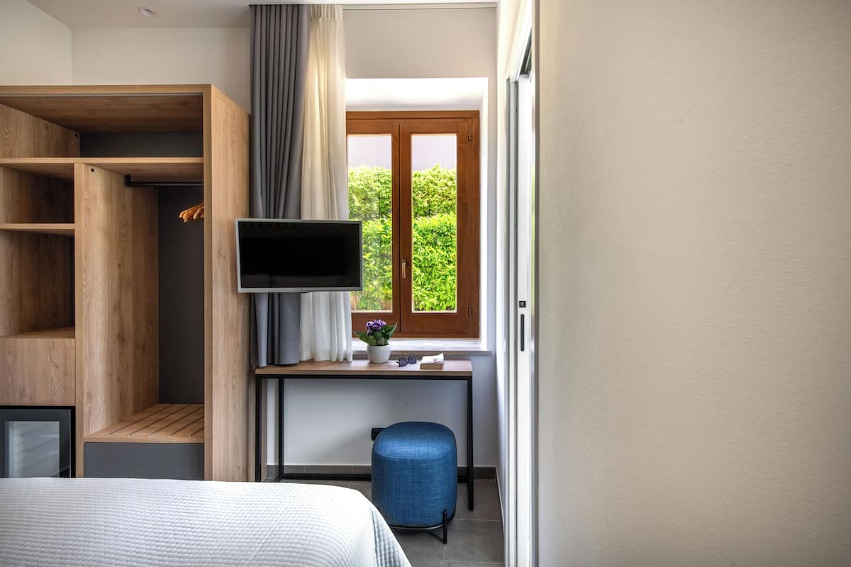A bedroom with a bed, a desk with a TV, a wardrobe, and a window with a view of greenery.