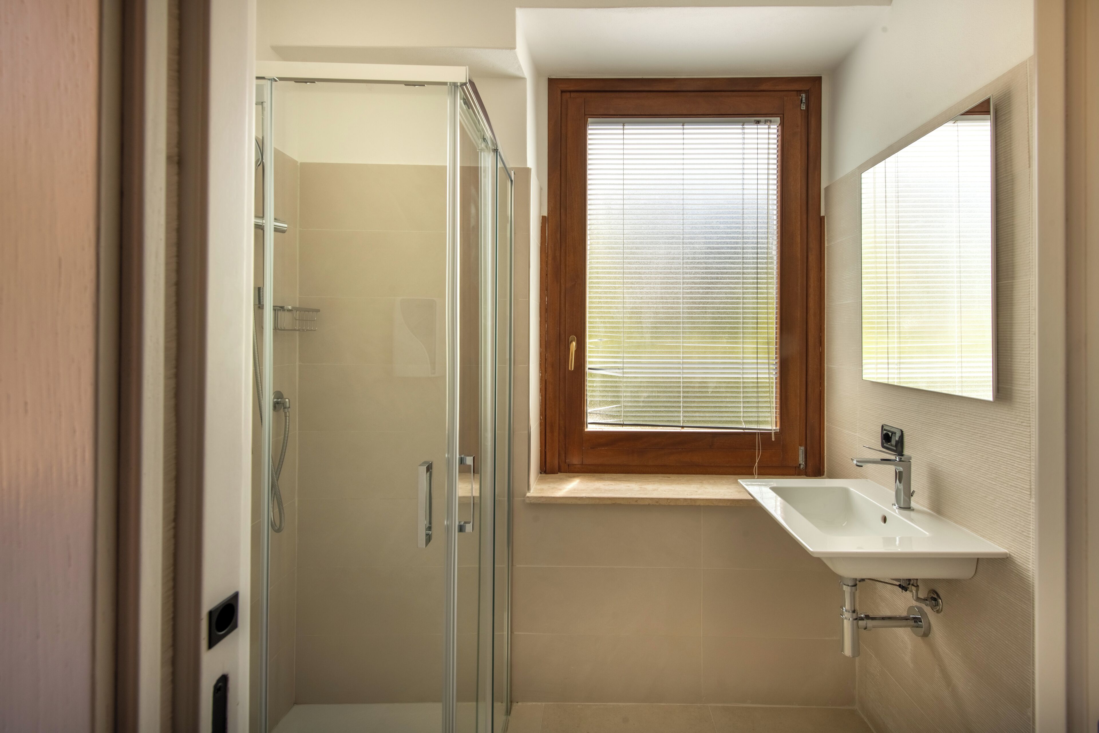 Un baño con cabina de ducha de vidrio, ventana con marco de madera y lavamanos blanco.