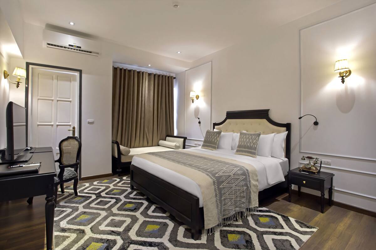 Ein Hotelzimmer mit einem großen Bett, einem Schreibtisch mit Computer, einem Sessel, einem Fernseher und einem gemusterten Teppich.