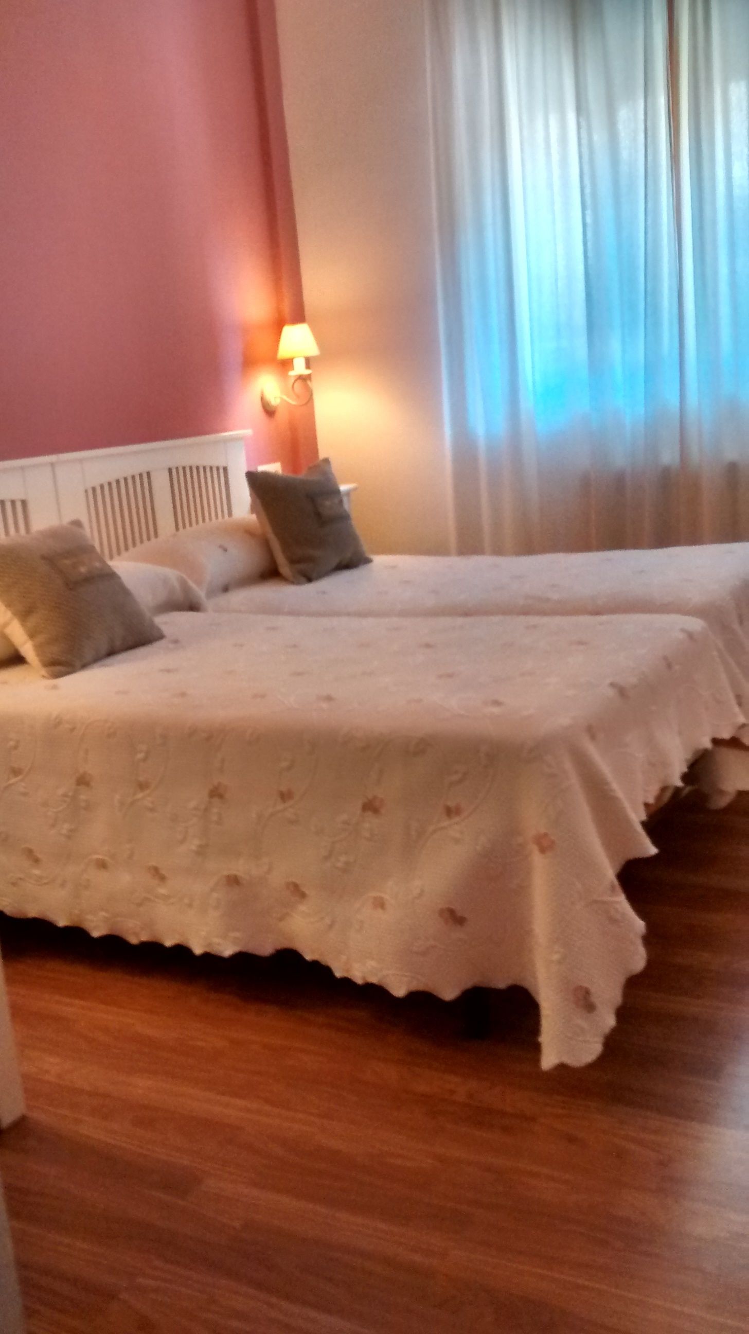 Una habitació d'hotel amb dos llits, un sòl de fusta, una làmpada de paret i una finestra amb cortines.