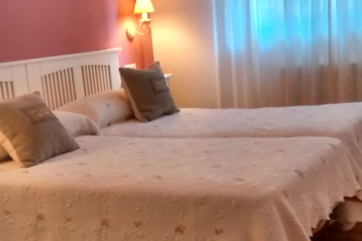 A hotel room with two beds, a wooden floor, a wall lamp, and a window with curtains.