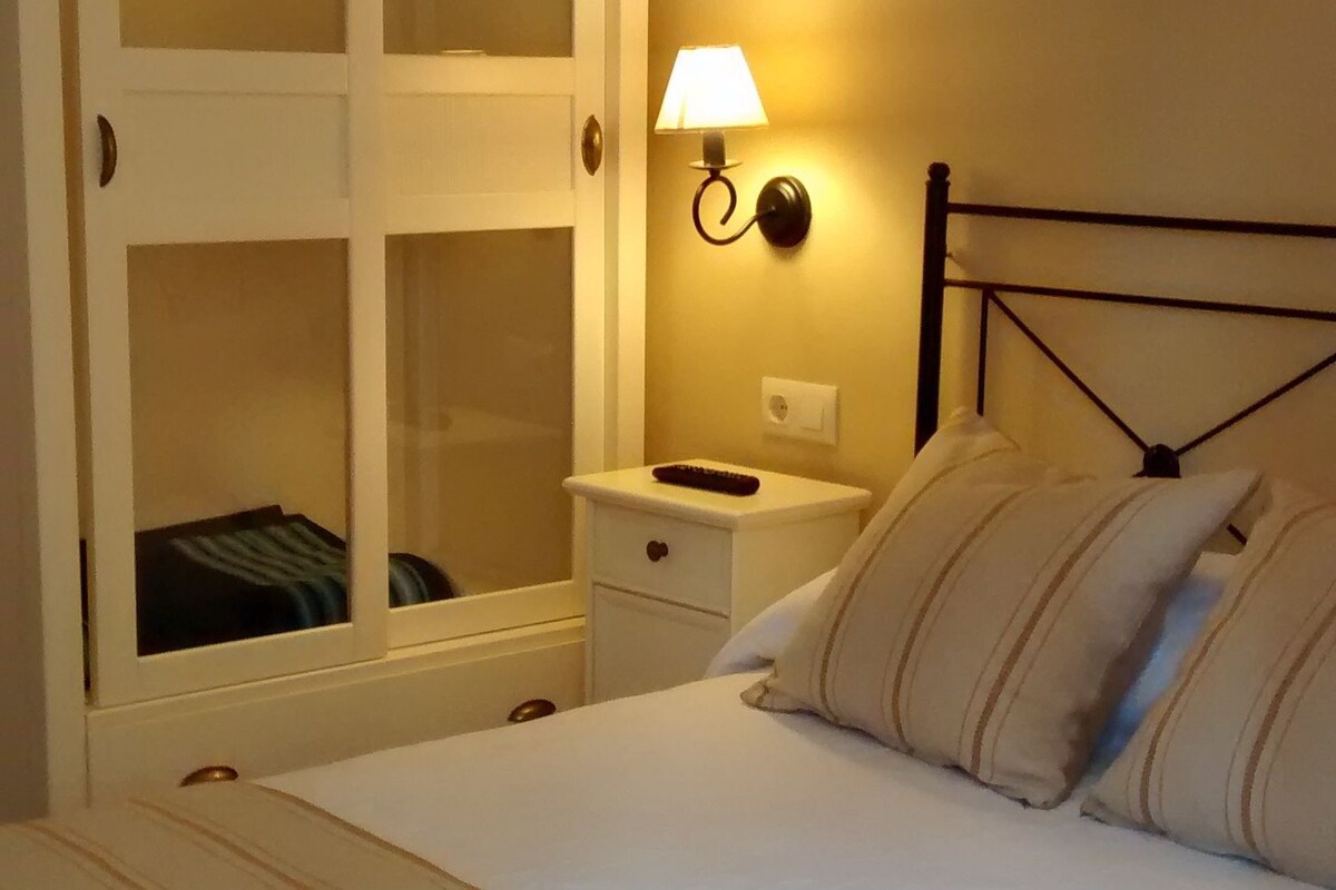 A bedroom with a bed, a bedside table, a nightstand, a wardrobe, and a wall-mounted lamp.
