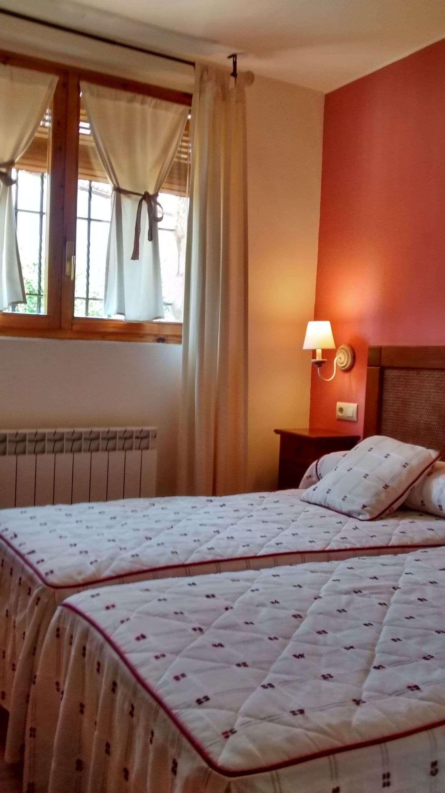 Habitación de hotel con dos camas, una ventana con cortinas, una lámpara de pared y un radiador.