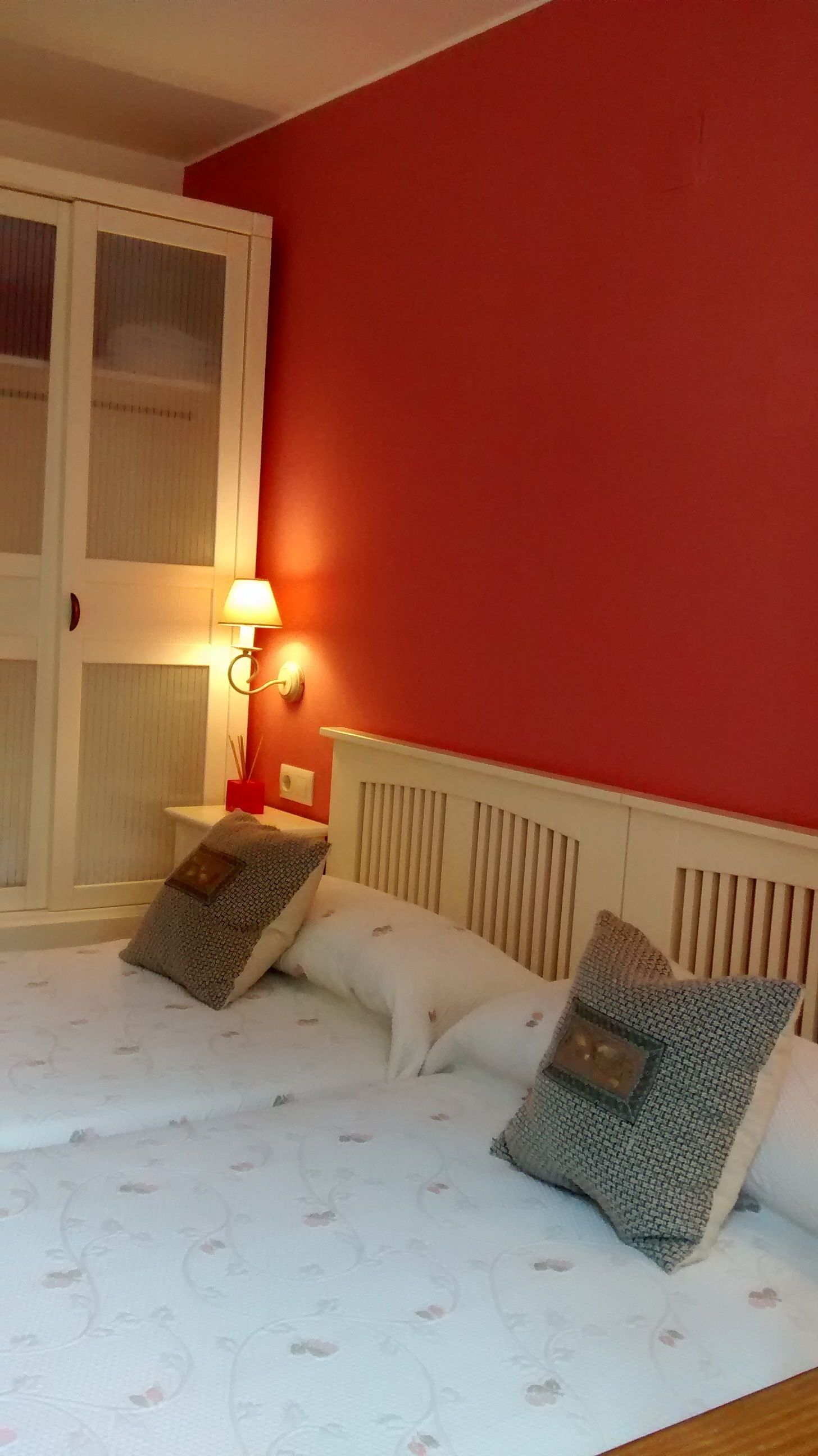 Un dormitorio con cama, almohadas, una mesita de noche, una lámpara y una ventana con persianas.
