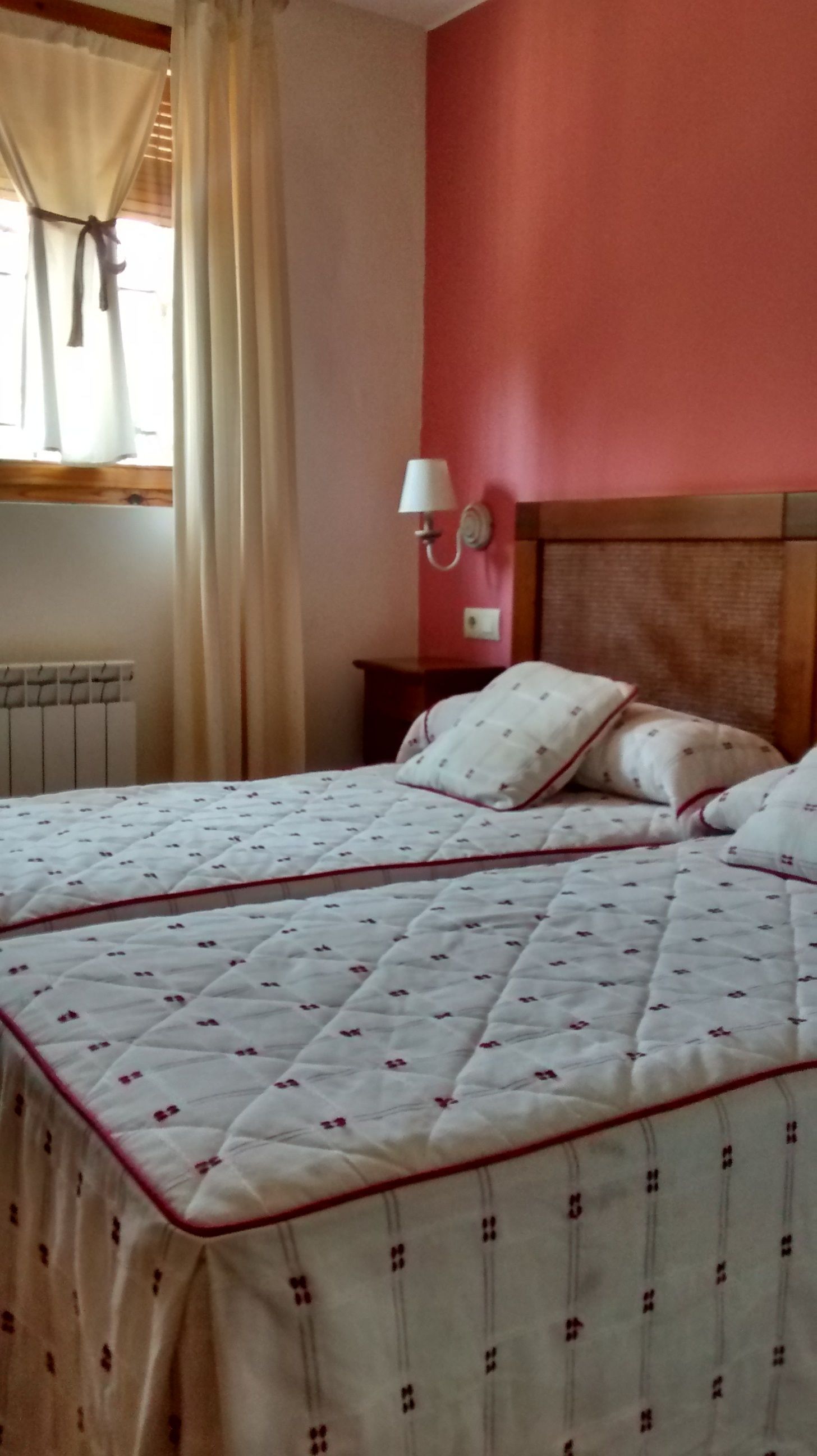 Un dormitorio con cama, mesita de noche, lámpara de pared y ventana con cortinas.