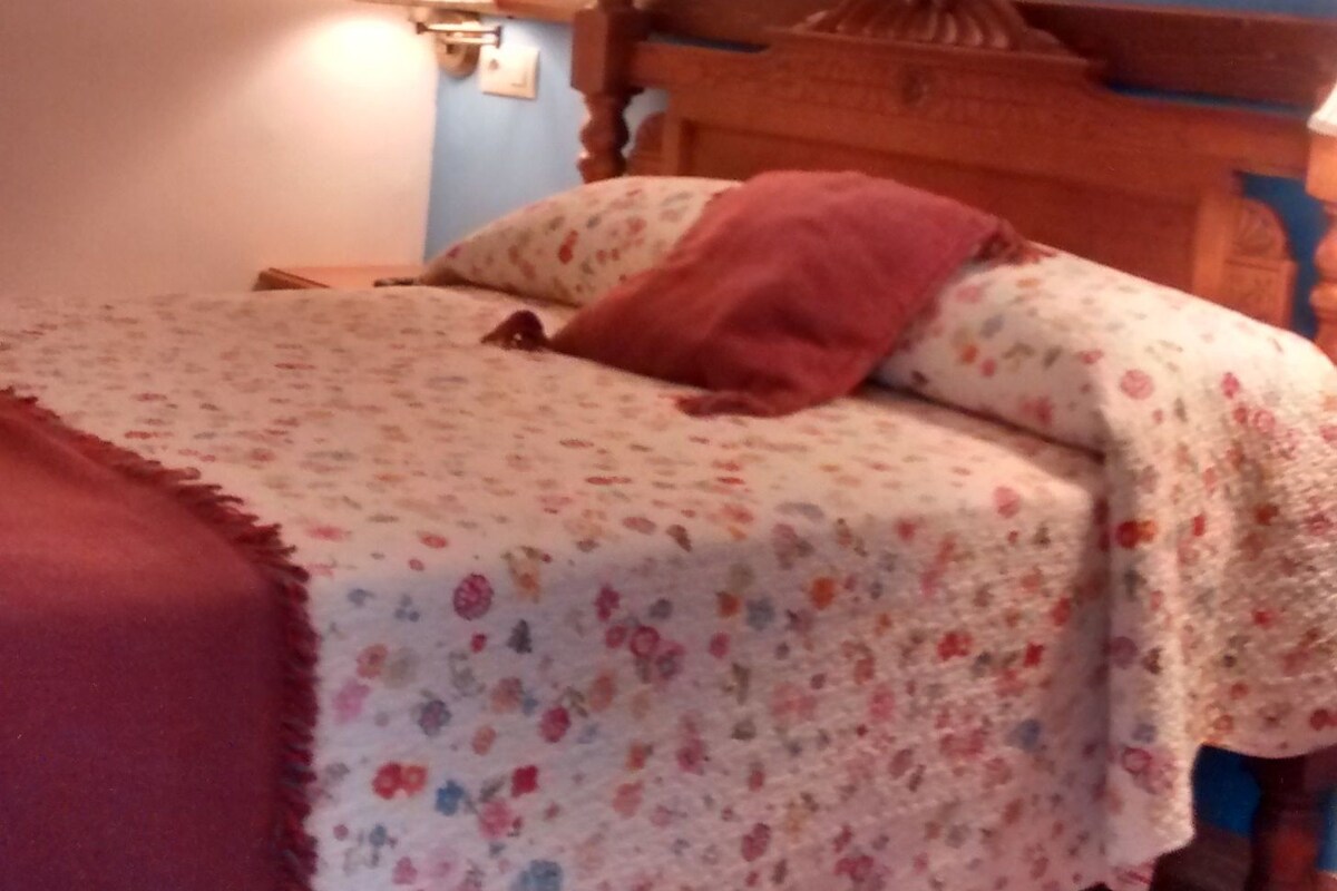A bedroom with a wooden bed, floral bedding, a red blanket, a wall-mounted lamp, and a window with a white curtain.