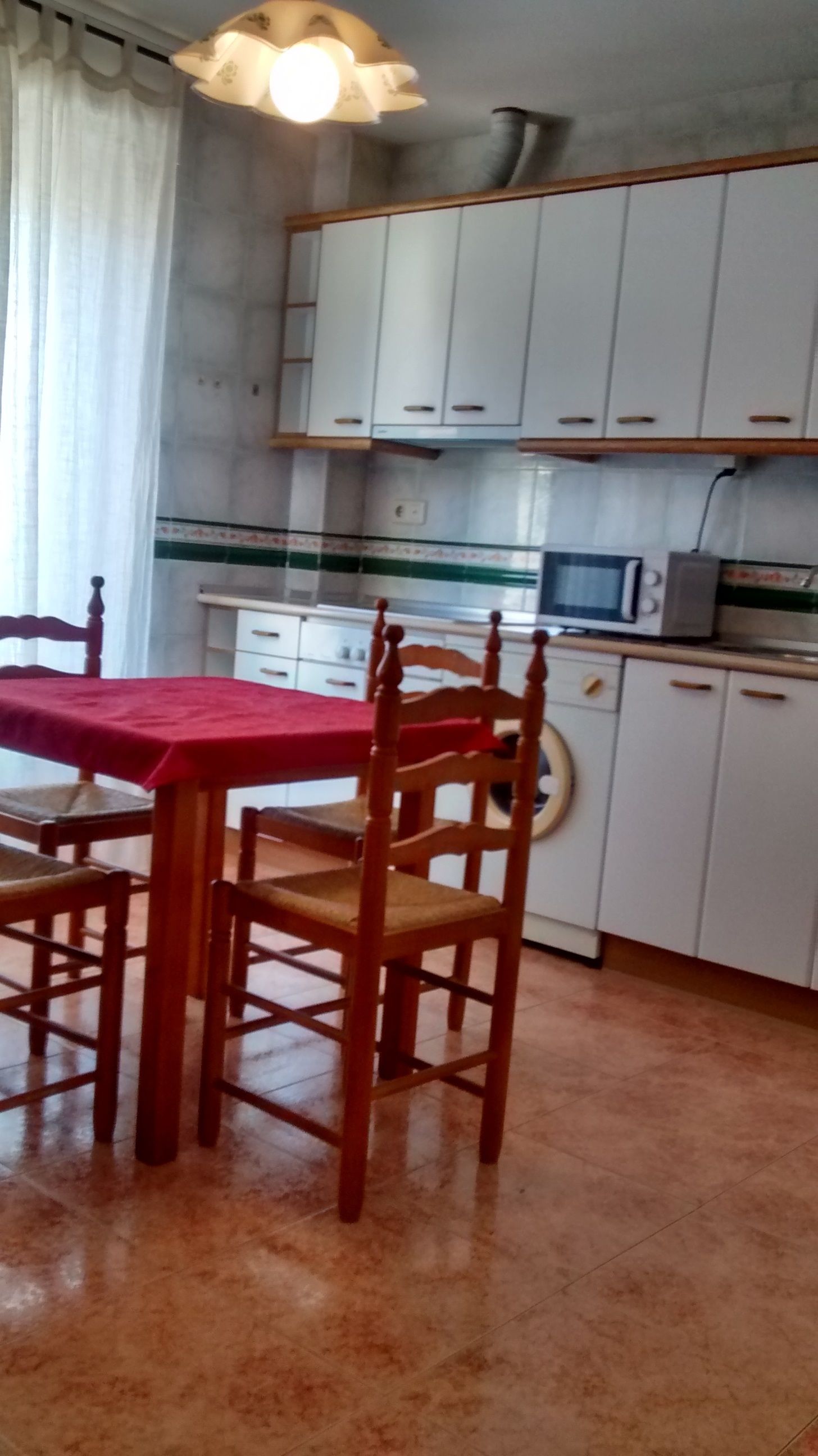 Une cuisine avec des meubles blancs, un four à micro-ondes et une table recouverte d’une nappe rouge, ainsi que des chaises en bois.