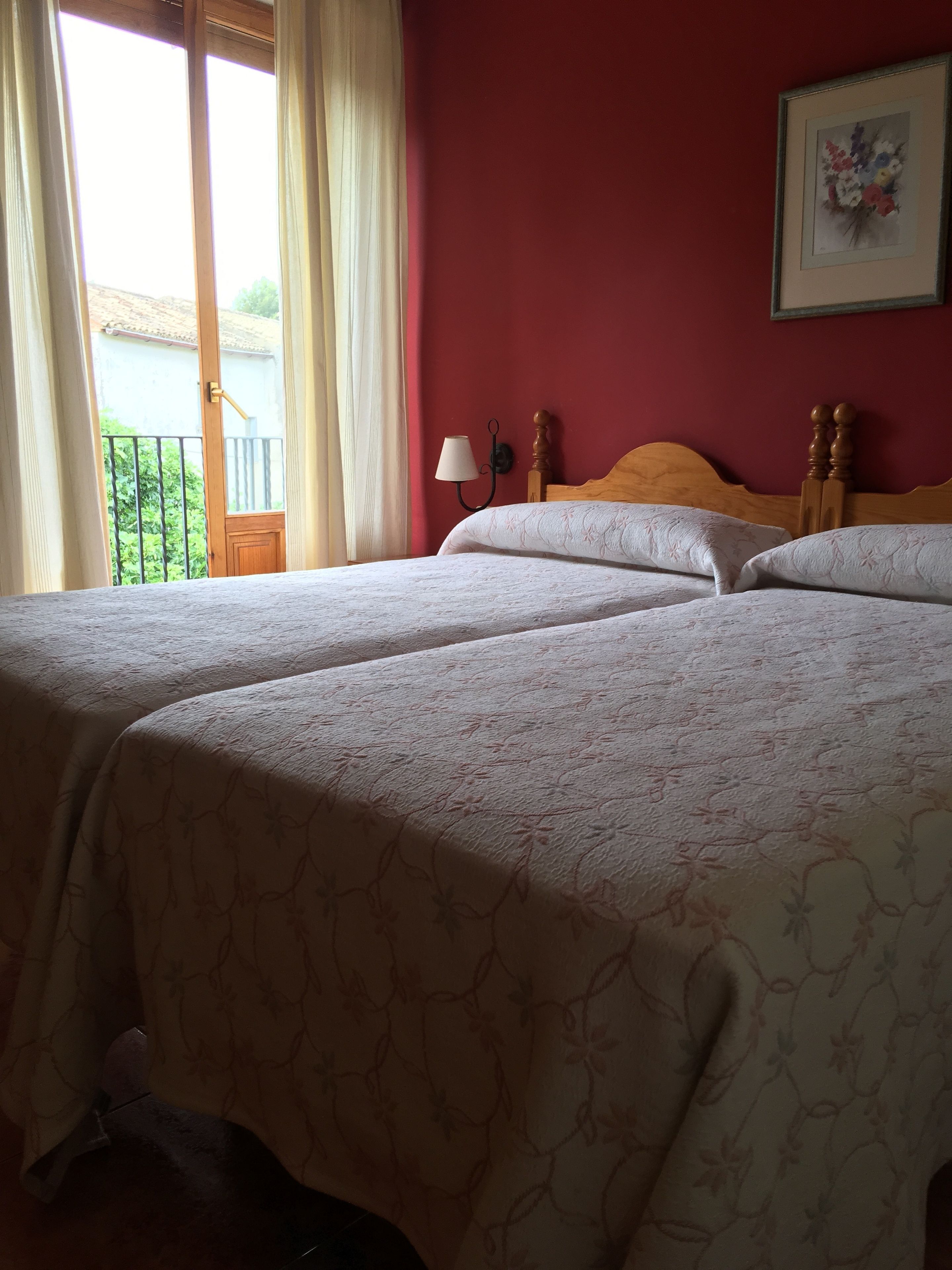 A hotel room with two beds, a wooden headboard, a wall-mounted lamp, and a view of greenery outside.