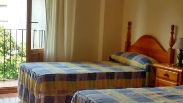 A room with two beds, a wooden headboard, a nightstand, a lamp, and a view of greenery outside.