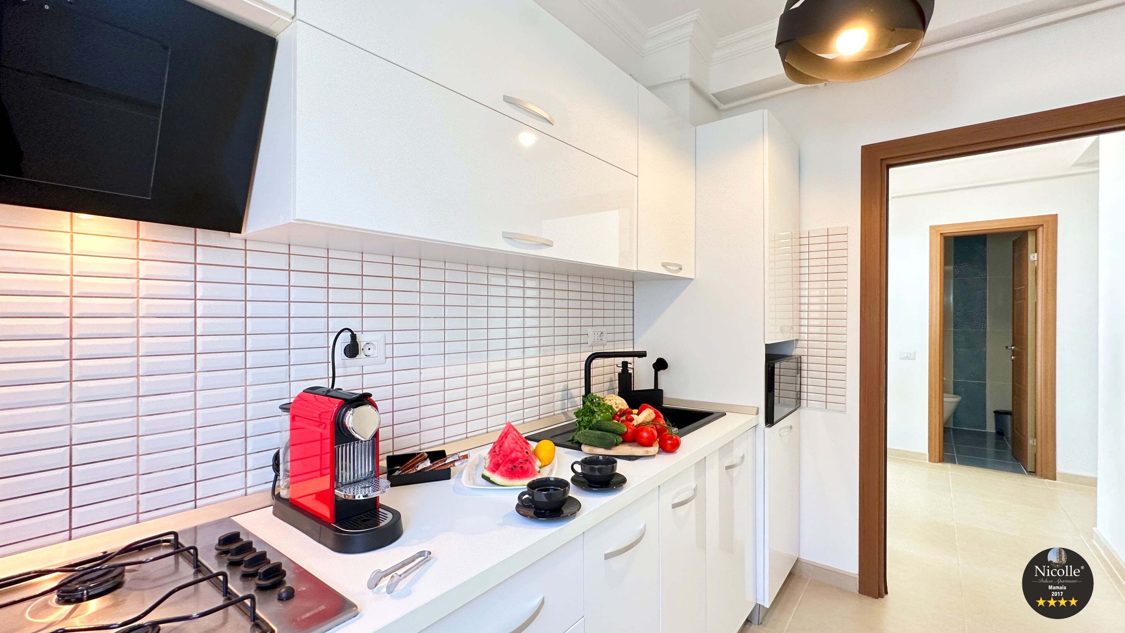 A modern kitchen with white cabinets, a black stove, and a red coffee maker. There are fresh vegetables on the counter and a sink with a black faucet.