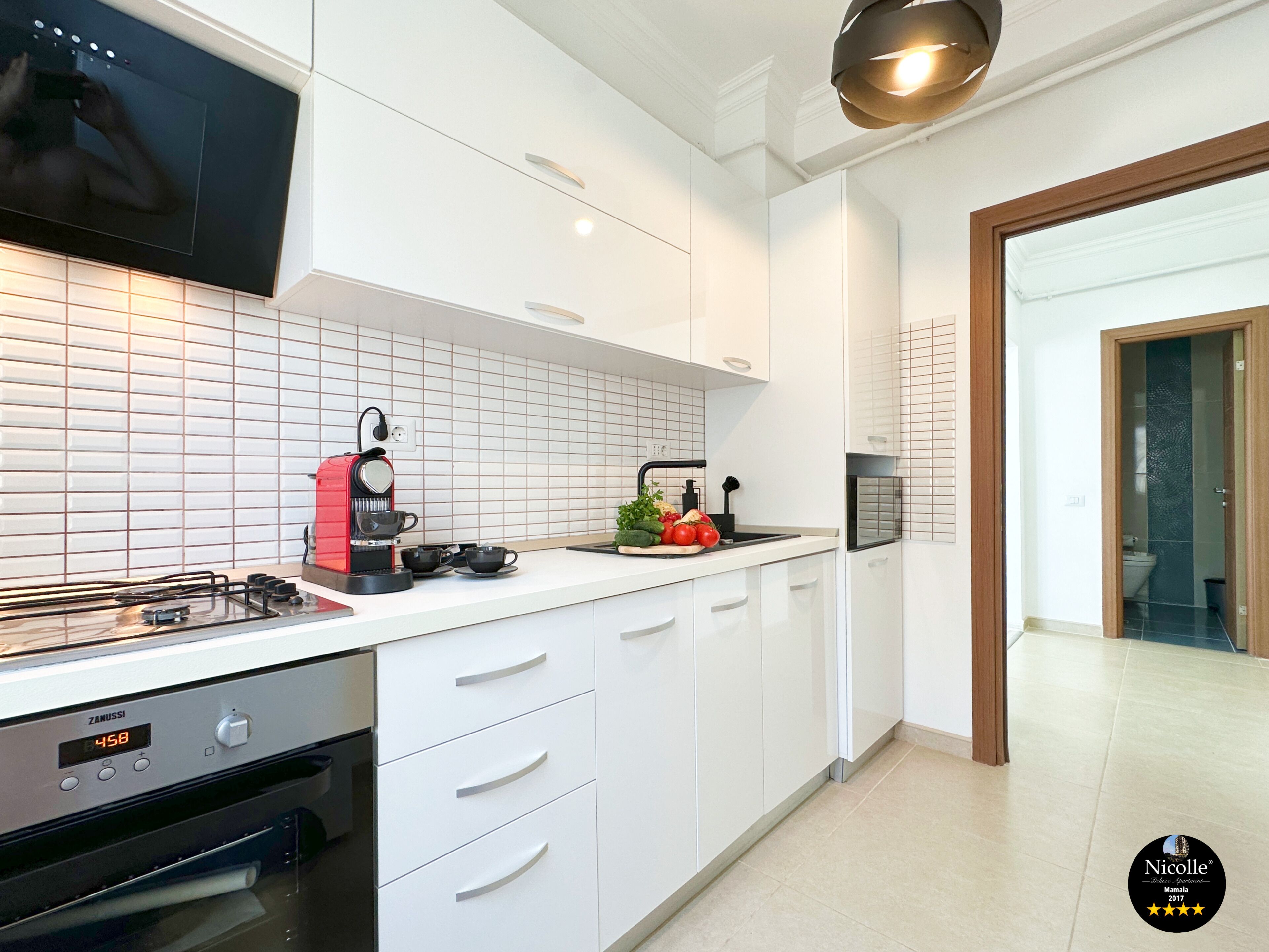 A modern kitchen with white cabinetry, a built-in oven, a coffee machine, and a large flat-screen TV mounted above the counter.