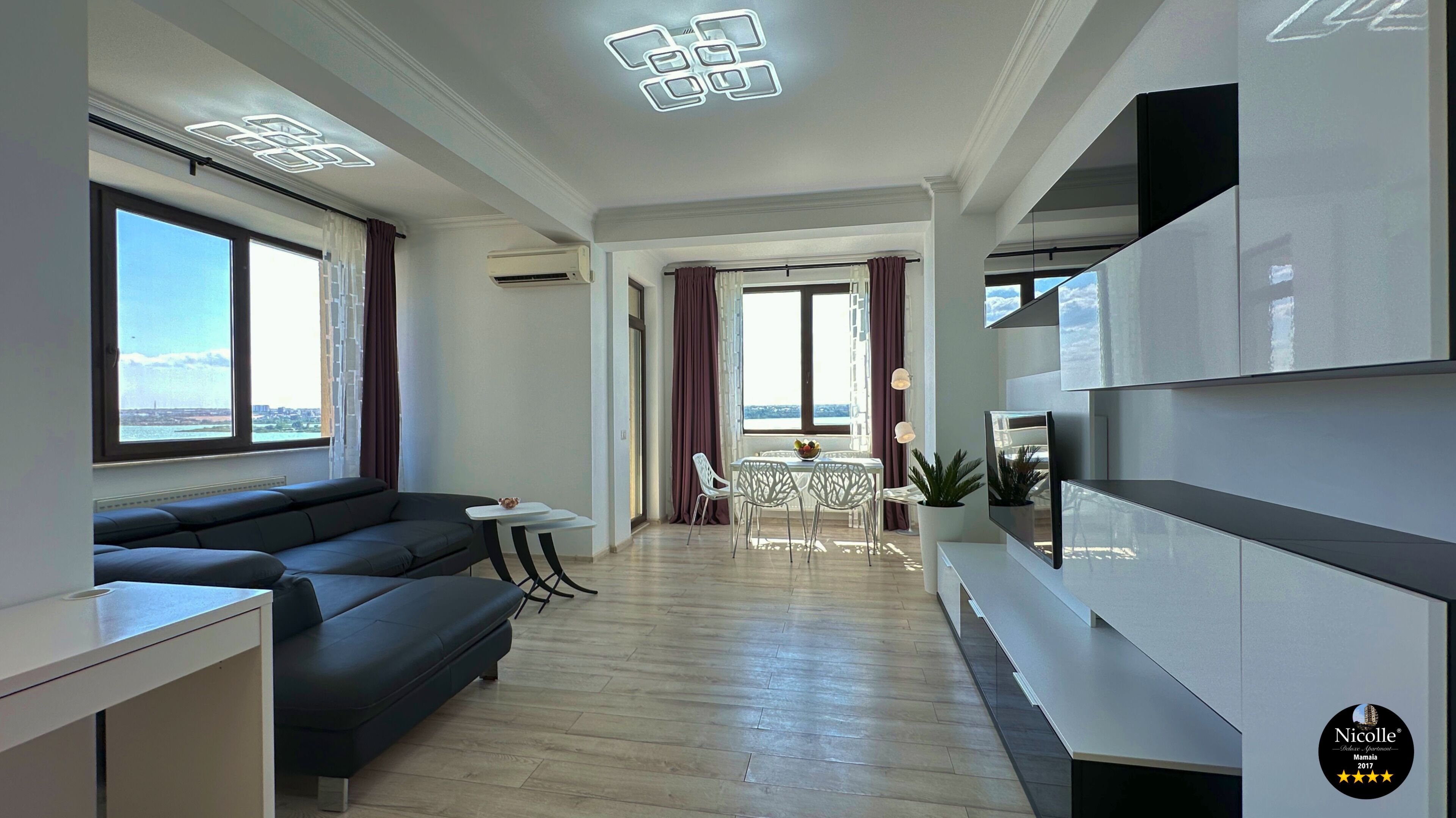 A modern living room with a sofa, dining table, and chairs, large windows with curtains, and a flat-screen TV mounted on the wall.