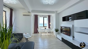Una sala de estar moderna con un sofá, una mesa de comedor con sillas, un televisor montado en la pared y vista al mar por la ventana.