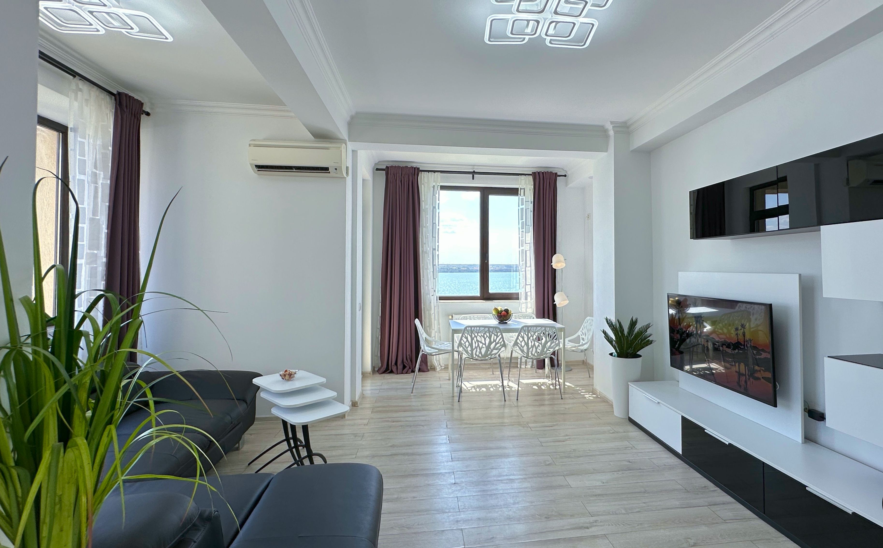 Een moderne woonkamer met een bank, een eettafel met stoelen, een aan de muur bevestigde televisie en uitzicht op de zee.