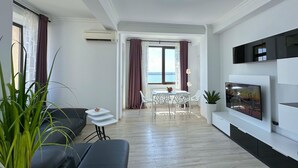 A modern living room with a sofa, a dining table with chairs, a TV mounted on the wall, and a view of the sea through the window. - Deluxe Nicolle Solid Residence Mamaia (Constanta)