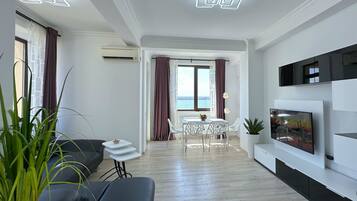 A modern living room with a sofa, a dining table with chairs, a TV mounted on the wall, and a view of the sea through the window.