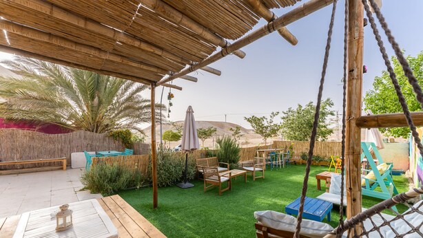 Terraza de madera con mesa y sillas, rodeada por un jardín vallado con palmeras y una zona de juegos.