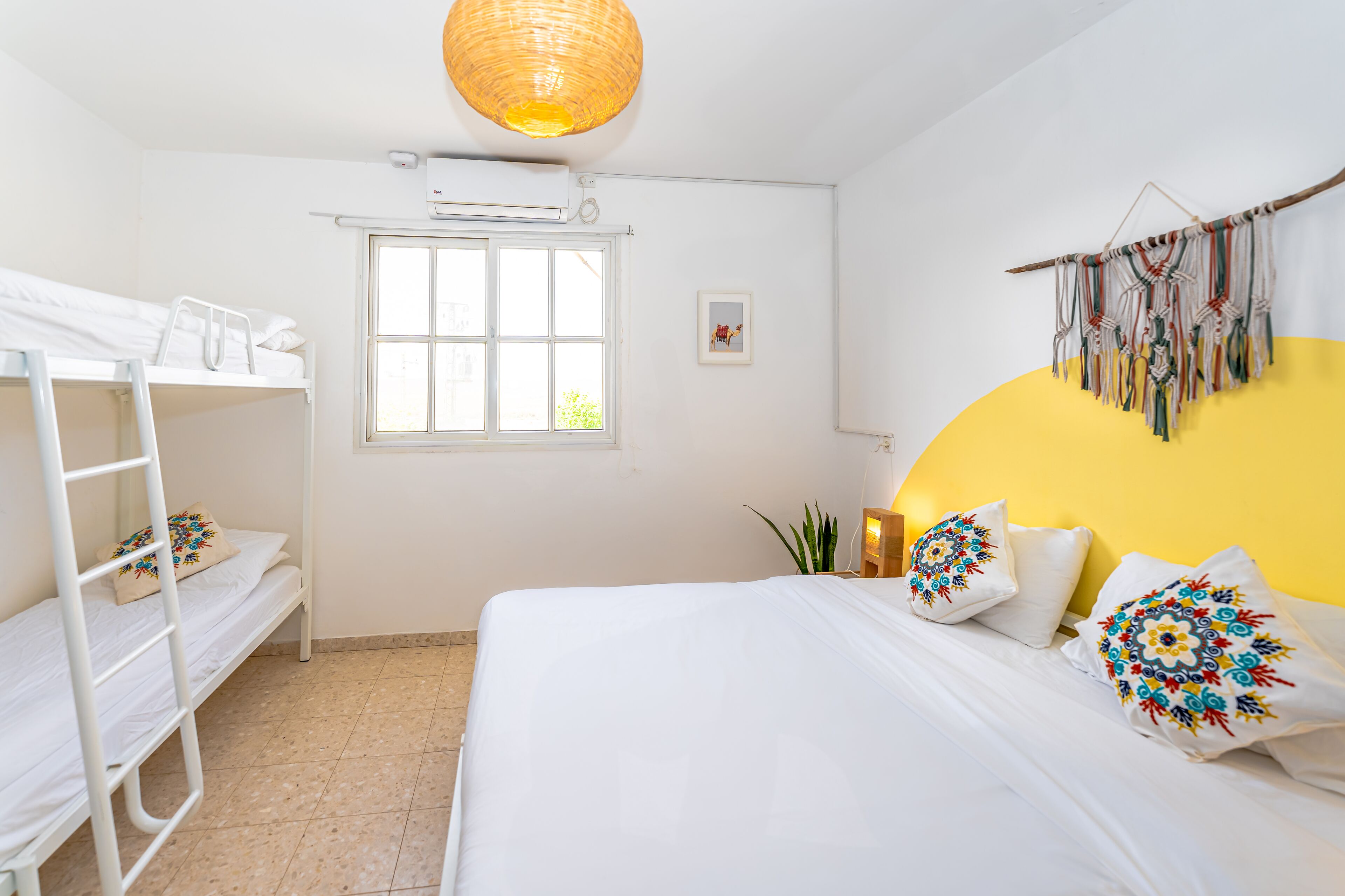 A room with a bunk bed, a single bed with colorful pillows, a hanging macrame wall decoration, and a window providing natural light.