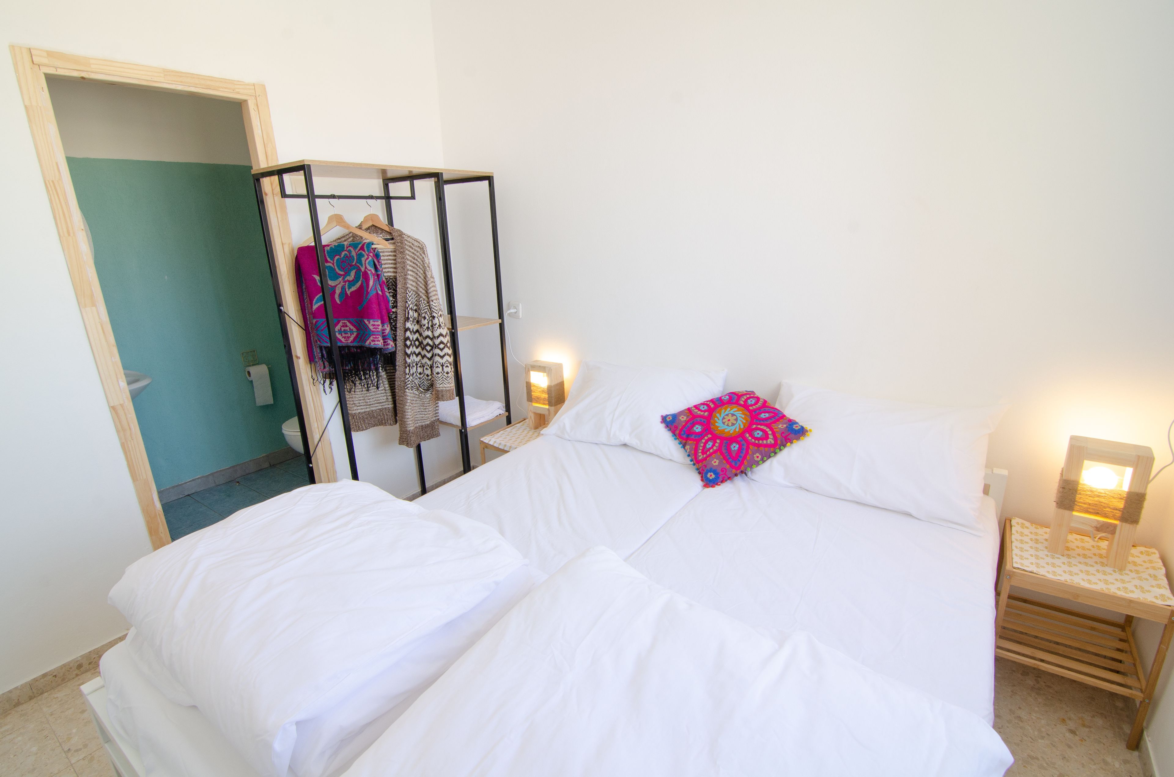 Un dormitorio con cama, mesita de noche, perchero y un baño visible a través de una puerta abierta.